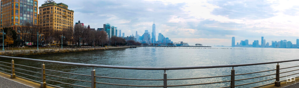 Panoramic view of a waterfront promenade with a metal railing, orange brick buildings on the left and a distant city skyline across the water under a cloudy sky.
