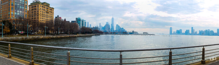 Panoramic view of a waterfront promenade with a metal railing, orange brick buildings on the left and a distant city skyline across the water under a cloudy sky.