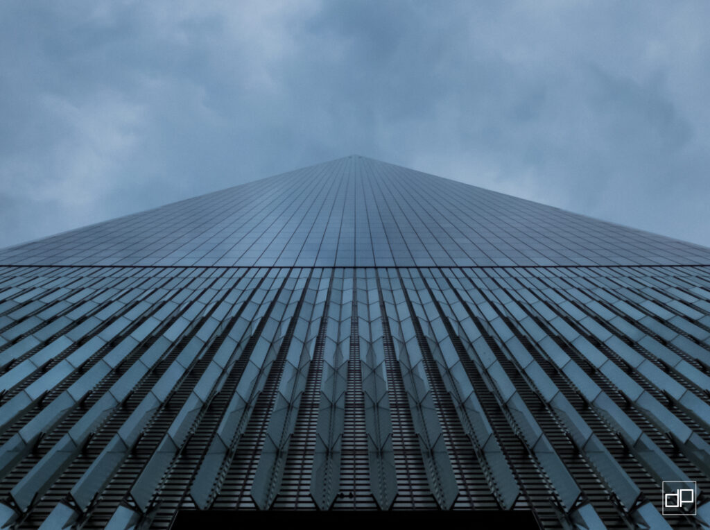 Low-angle view of a blue-tinted glass skyscraper converging to a sharp apex against a cloudy sky, with a white watermark in the bottom-right corner.