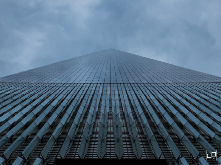 Low-angle view of a blue-tinted glass skyscraper converging to a sharp apex against a cloudy sky, with a white watermark in the bottom-right corner.