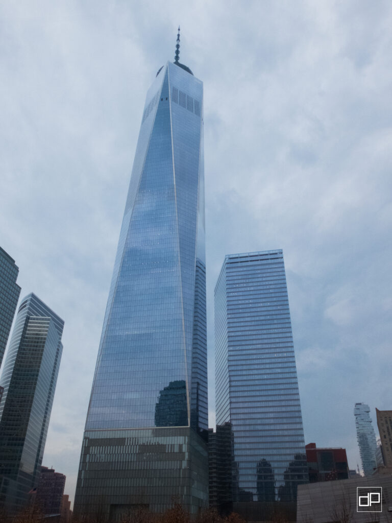 Tall glass skyscrapers in a cityscape under a cloudy sky, with One World Trade Center-like tower towering above neighboring buildings.