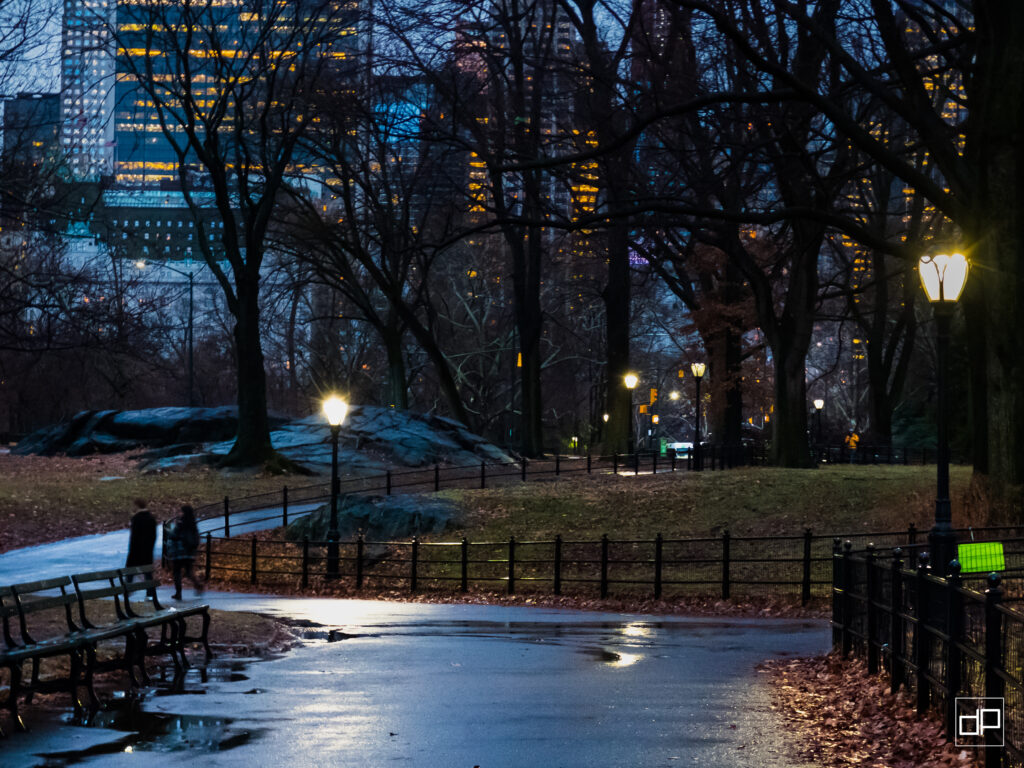 Evening park path with wet pavement, bare trees, benches, and glowing streetlamps; city skyline lit in the distance after rain.