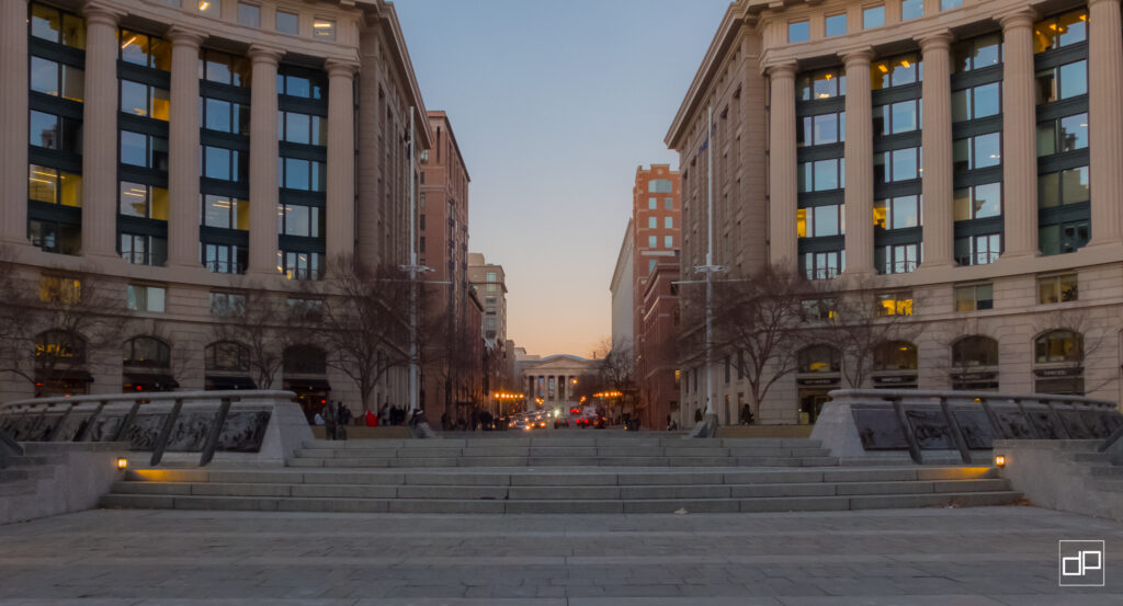 Symmetrical city plaza between tall beige buildings with columns, steps down to a street at dusk; faint pedestrians visible.