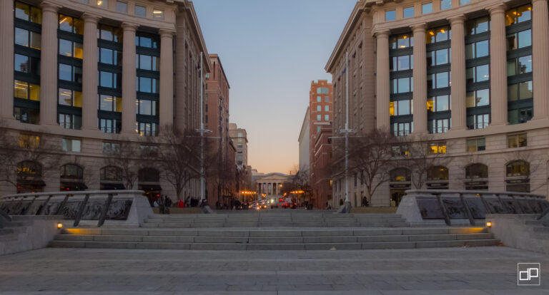 Symmetrical city plaza between tall beige buildings with columns, steps down to a street at dusk; faint pedestrians visible.