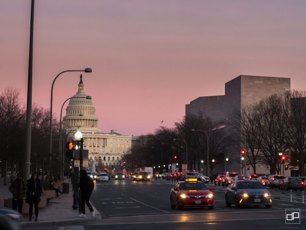 Capitol dome visible on a city street at dusk with cars and pedestrians and a pink sky behind leafless trees.