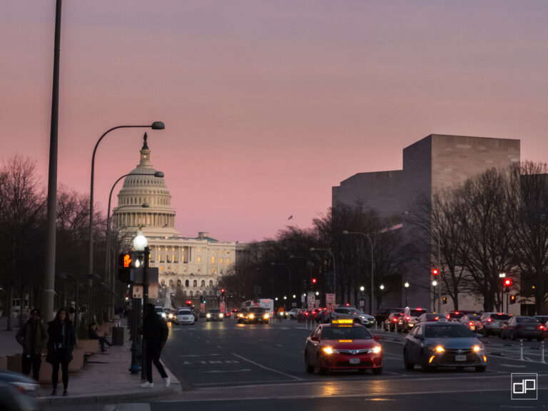 Capitol dome visible on a city street at dusk with cars and pedestrians and a pink sky behind leafless trees.