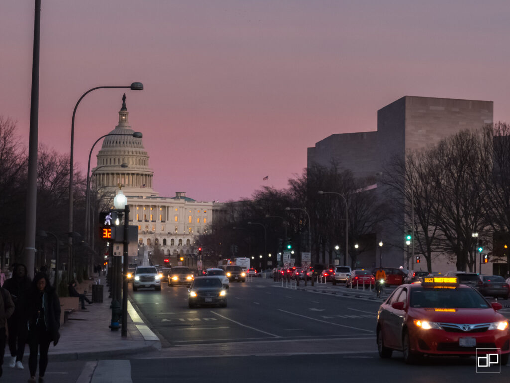 Capitol building at dusk visible along a busy city street with cars, pedestrians, and a pink sunset sky in the background