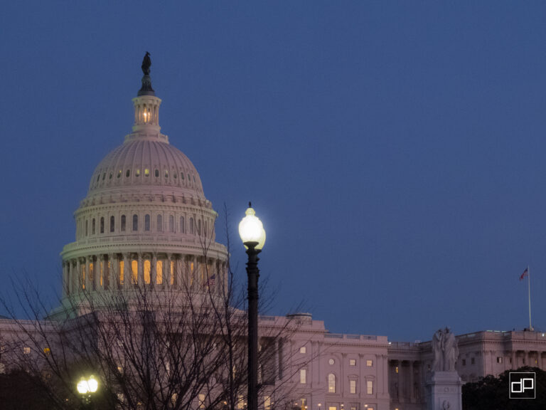 Dusk view of the U.S. Capitol dome with statue atop, lit windows, and a street lamp in the foreground against a blue sky.