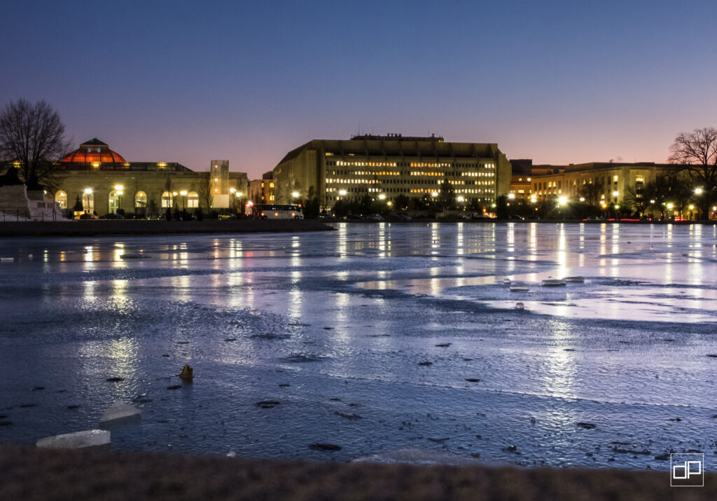 City waterfront at dusk with illuminated buildings reflecting on a frozen river/surface of ice.