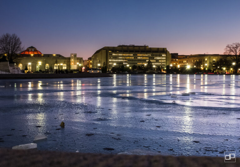 City waterfront at dusk with illuminated buildings reflecting on a frozen river/surface of ice.