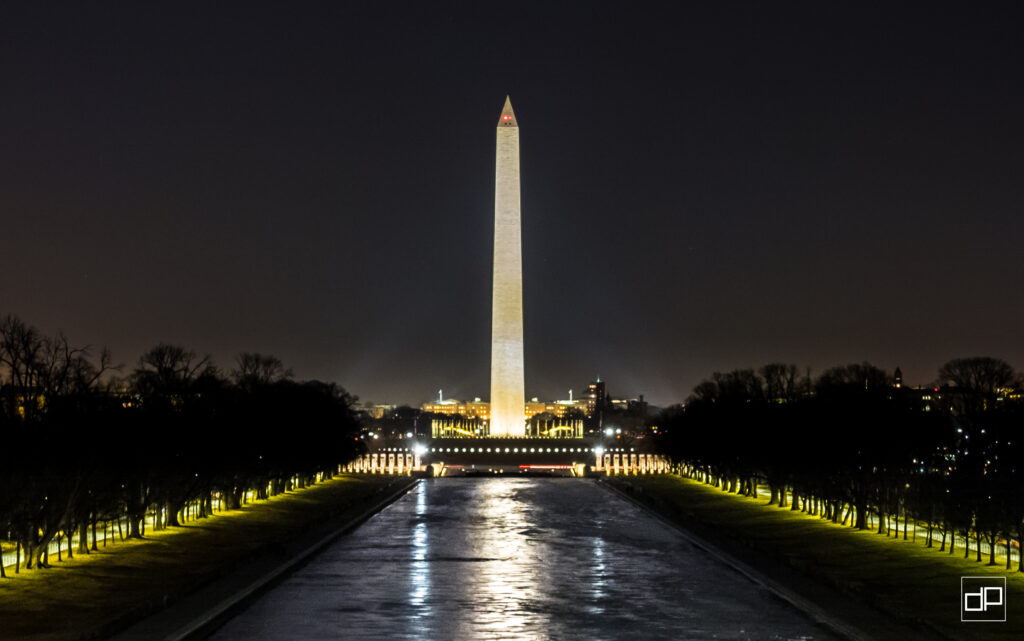 Night view of the Washington Monument illuminated, with the reflecting pool and lit pathways in the foreground and leafless trees lining the sides at dusk.