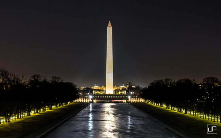 Night view of the Washington Monument illuminated, with the reflecting pool and lit pathways in the foreground and leafless trees lining the sides at dusk.