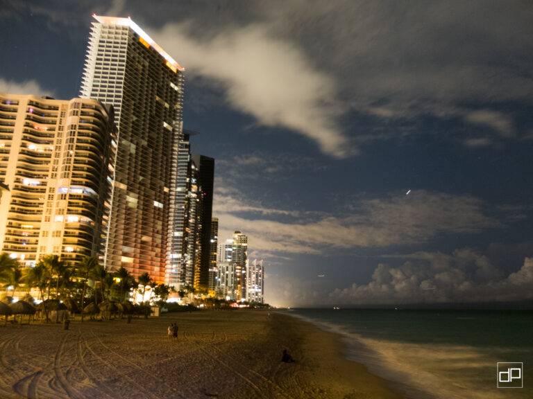 Nighttime beach scene with illuminated high-rise buildings along the shore, palm trees in the foreground, and a calm ocean to the right. A couple walks on the sand near the water under a cloudy sky.
