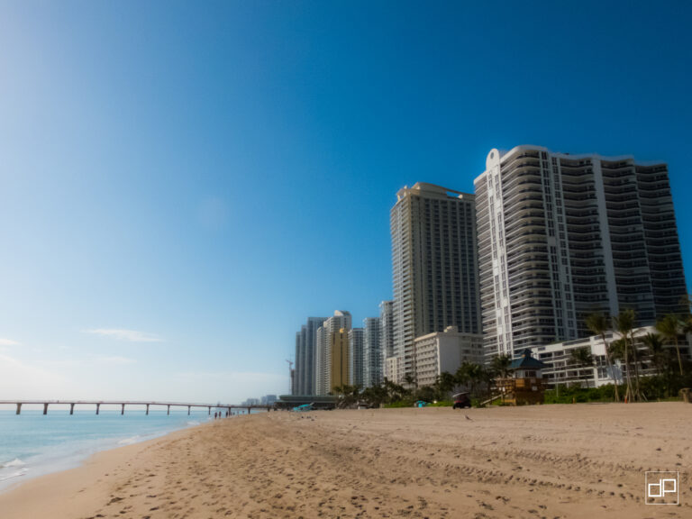 Sunny beach with high-rise buildings on the right and a pier extending into calm water under a clear blue sky.