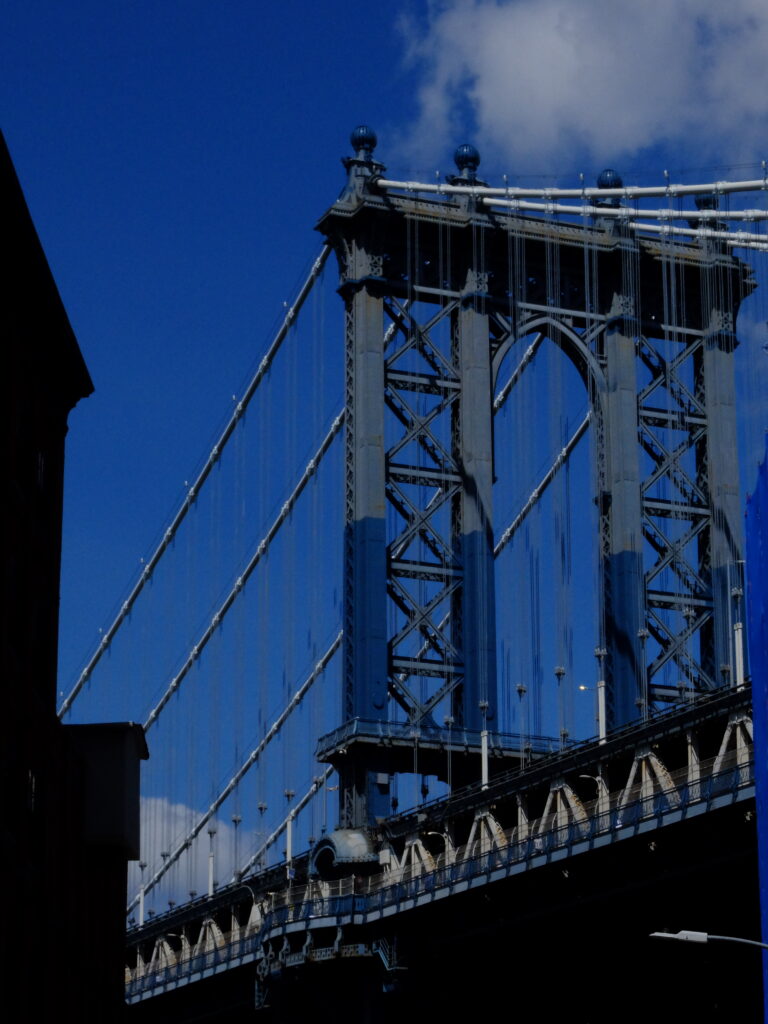 Close-up of a towering steel suspension bridge with vertical cables against a bright blue sky, highlighting the structure's metalwork.