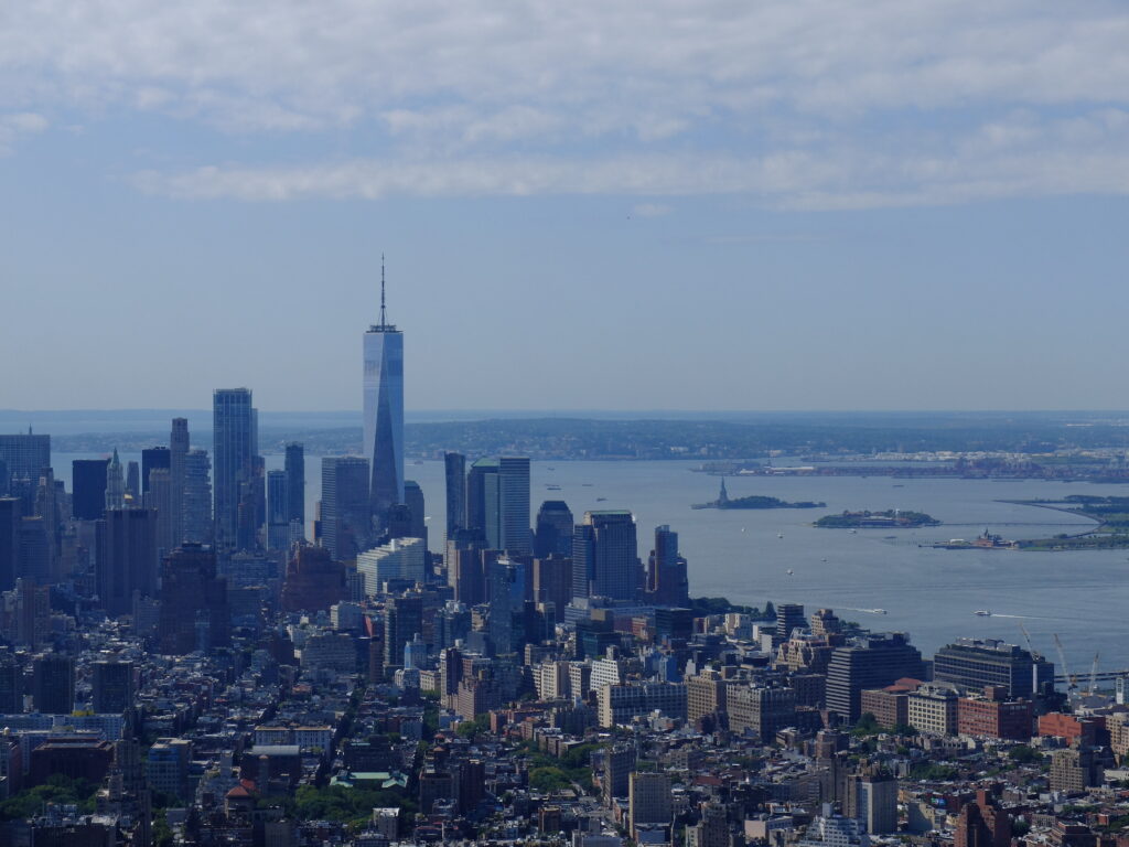 Aerial cityscape of Manhattan with the One World Trade Center rising above other buildings near the river under a blue sky.