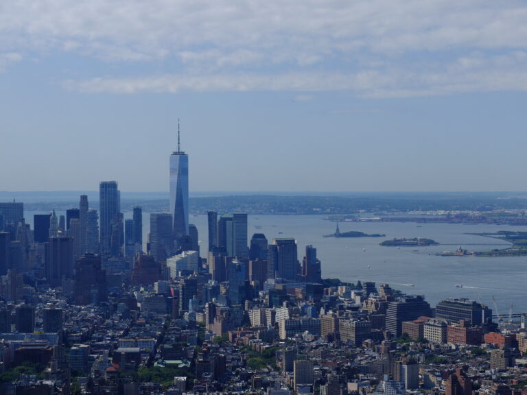 Aerial cityscape of Manhattan with the One World Trade Center rising above other buildings near the river under a blue sky.