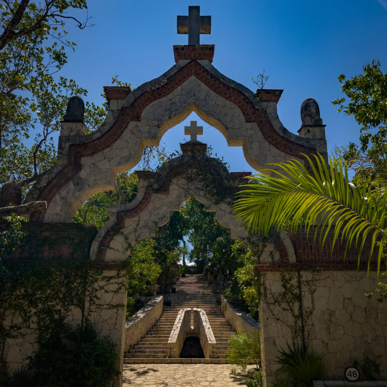 Stone archway and central staircase flanked by stone walls, topped with a cross against a bright blue sky, surrounded by greenery.
