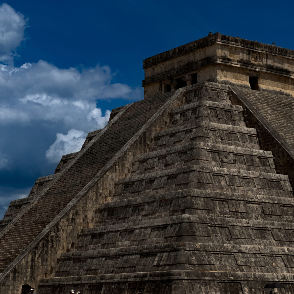 Stone stepped pyramid structure against a deep blue sky with clouds; a small group of tourists at the bottom left.