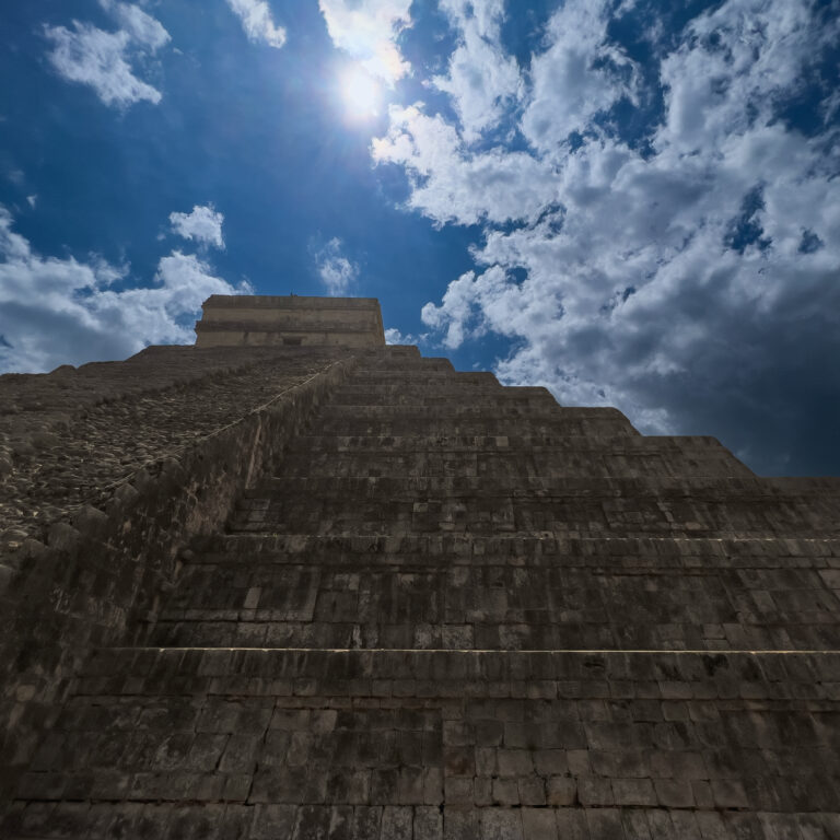 Stone pyramid stairs ascending toward a bright blue sky with a sun flare above.