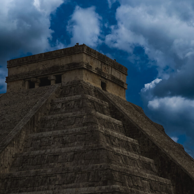 Top of an ancient stepped stone pyramid against a dramatic blue, cloudy sky.