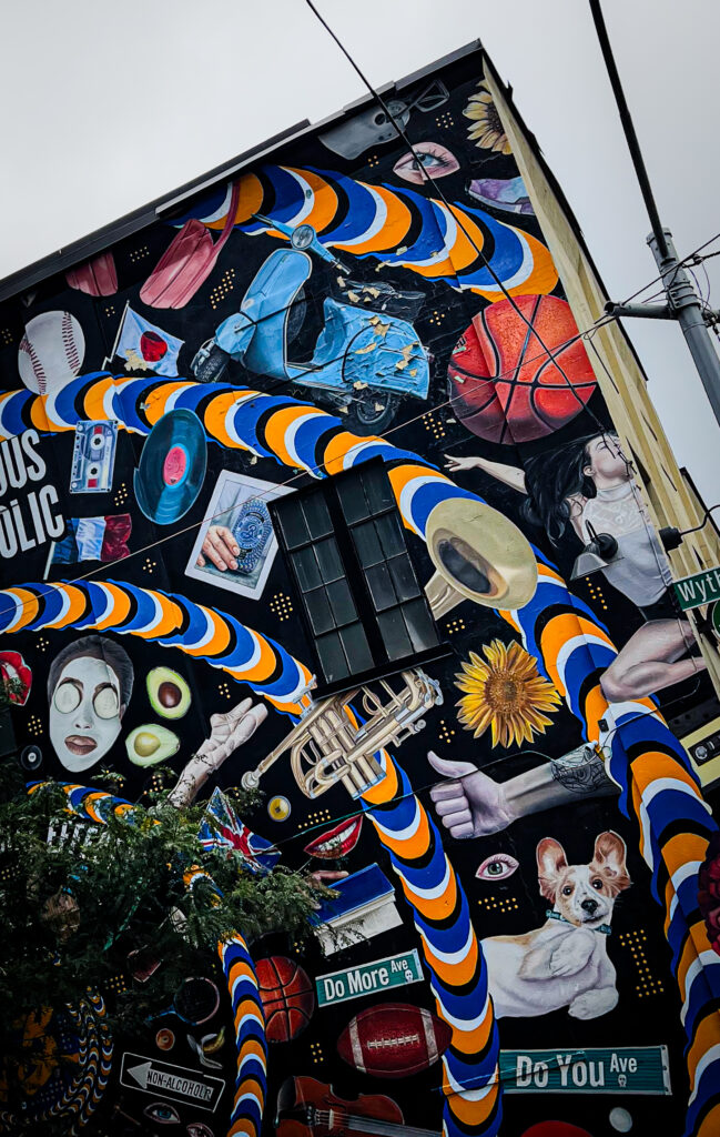 Vibrant mural on a building with blue and orange spirals, sports imagery (basketball, football), faces, a sunflower, and assorted collage elements.