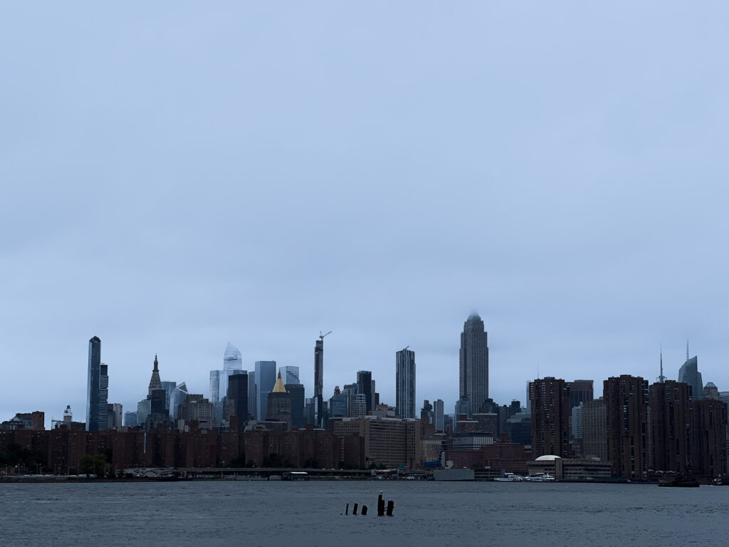 City skyline along a river with tall modern buildings under a pale blue cloudy sky, seen from the water's edge under overcast conditions.