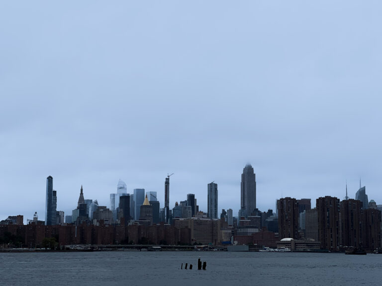 City skyline along a river with tall modern buildings under a pale blue cloudy sky, seen from the water's edge under overcast conditions.