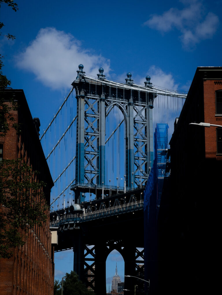 Brooklyn Bridge tower and suspension cables rise between brick buildings, with the Empire State Building visible in the distance against a blue sky.