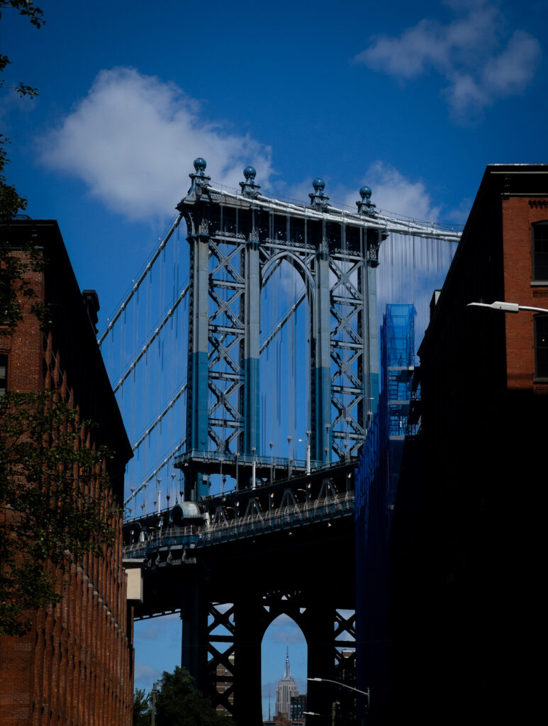Brooklyn Bridge tower and suspension cables rise between brick buildings, with the Empire State Building visible in the distance against a blue sky.