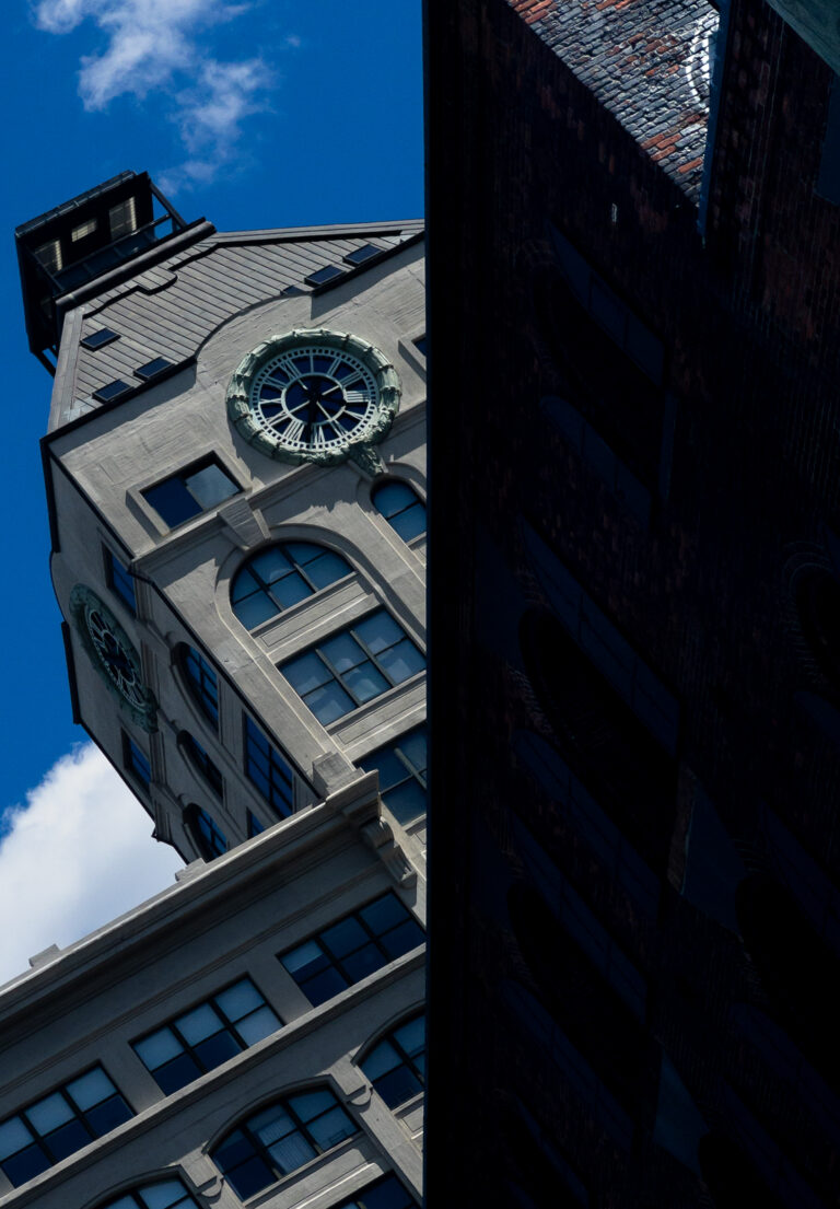 Two buildings viewed at an angle: a light-colored clock tower with arched windows against a blue sky on the left, and a dark, reflective brick facade on the right creating a clear split in the image.