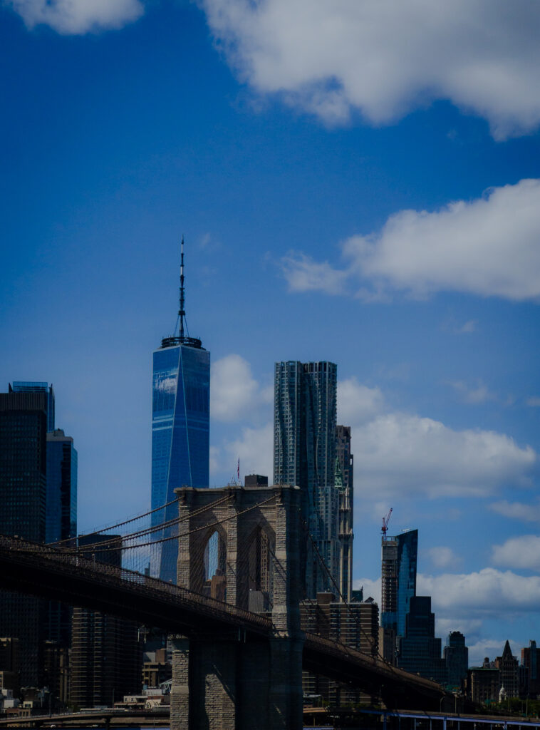 View of Manhattan skyline with One World Trade Center and Brooklyn Bridge in the foreground against a blue sky with clouds