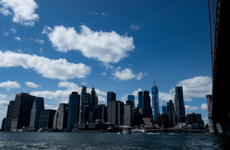 Wide view of the New York City skyline along the waterfront under a blue sky with white clouds.