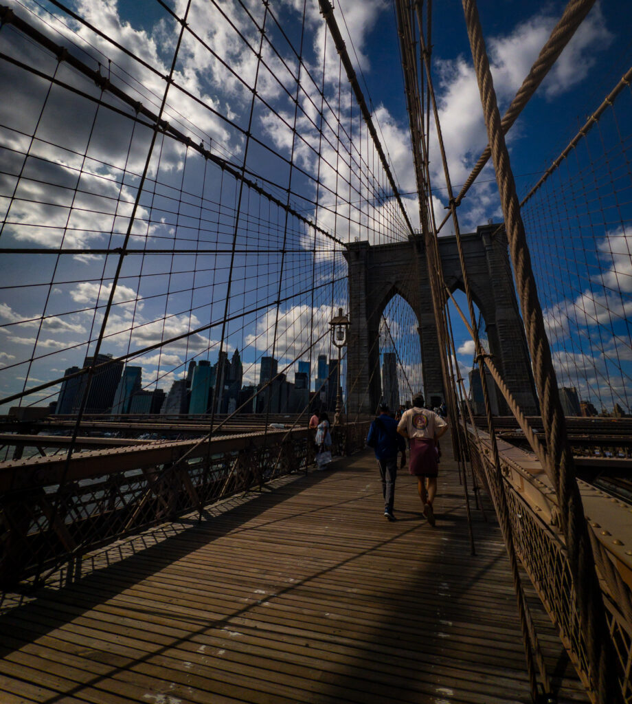 Pedestrians walk along the wooden path of the Brooklyn Bridge with crisscrossing steel cables and the NYC skyline in the distance under a blue sky.