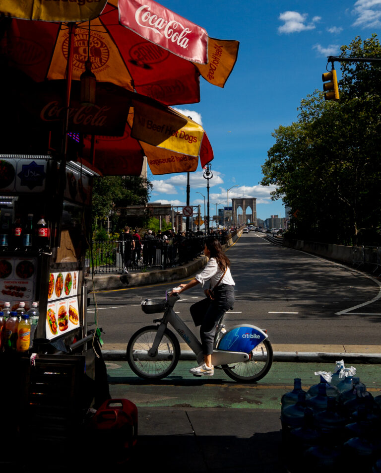 Street food cart under red Coca-Cola umbrellas on a sunny city street; a rider on a Citi Bike passes by with the Brooklyn Bridge in the distance under a blue sky.