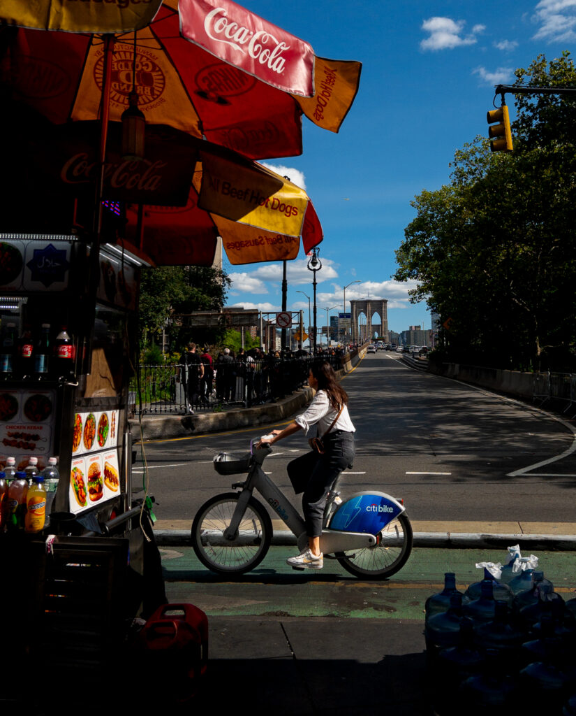 Street food cart under red Coca-Cola umbrellas on a sunny city street; a rider on a Citi Bike passes by with the Brooklyn Bridge in the distance under a blue sky.