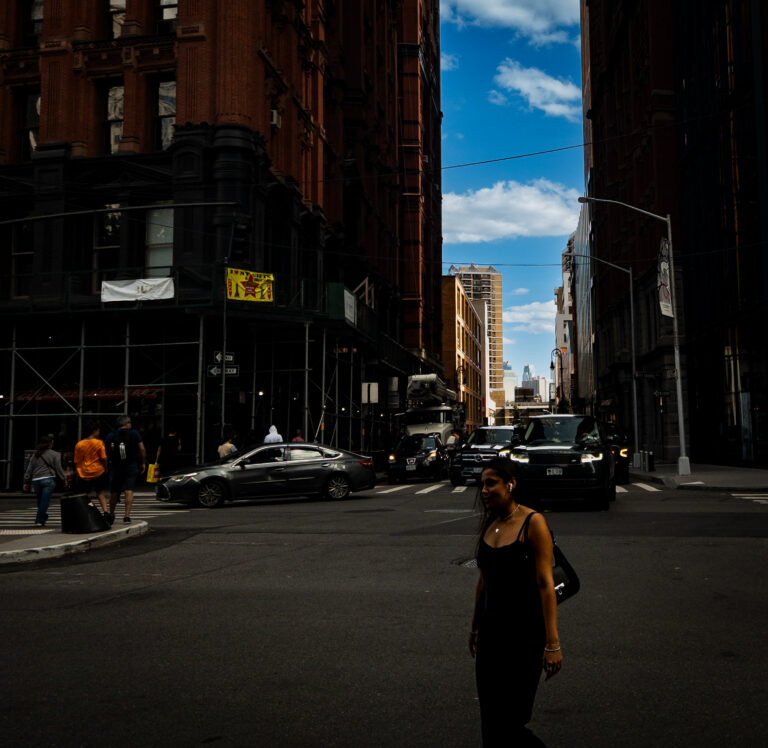Urban city street between tall brick buildings, blue sky visible above, with pedestrians and parked cars at a crosswalk edge.