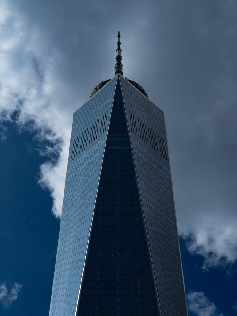 One World Trade Center skyscraper with a spire, glass facade, towering into a cloudy blue sky.