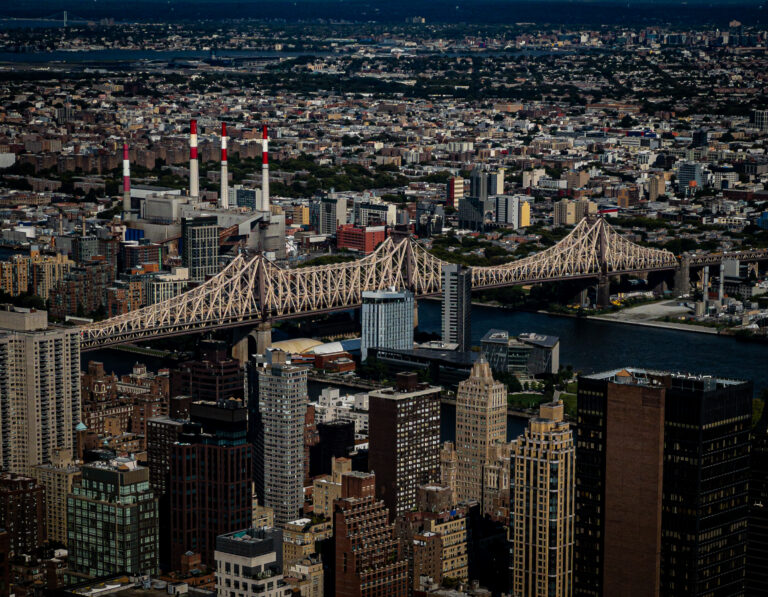 Wide cityscape showing a river, a large lattice bridge in the foreground, and dense high-rise buildings across the skyline.