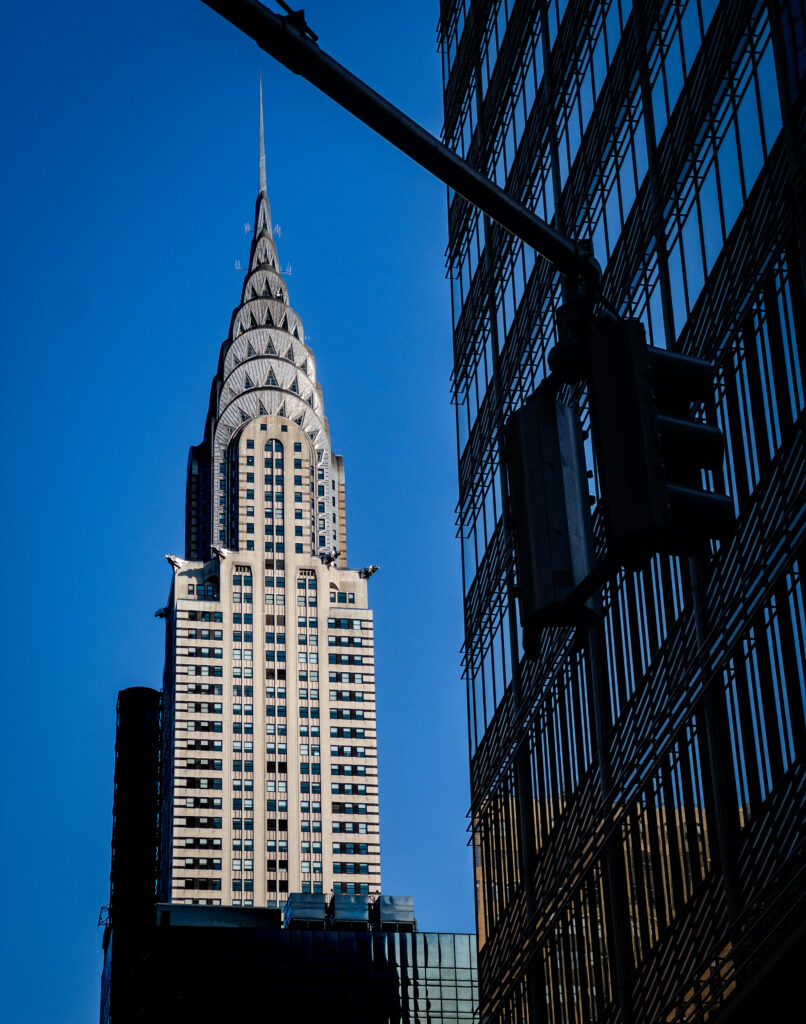 Chrysler Building with its spire rising beside a glass-clad office tower against a bright blue sky, showing architectural contrast.