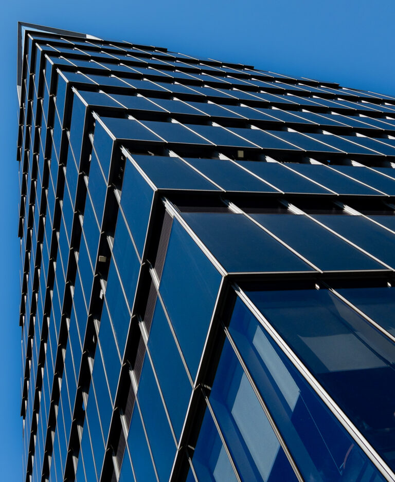 Modern glass office building with diagonal window panels reflecting sunlight against a clear blue sky