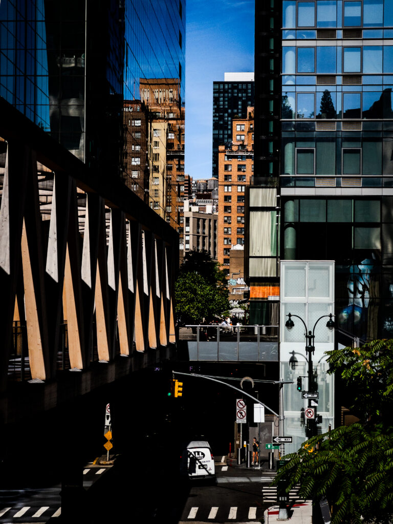 Urban street between glass-clad office buildings, with a diagonal wooden-support walkway on the left and a zebra crosswalk in the foreground.