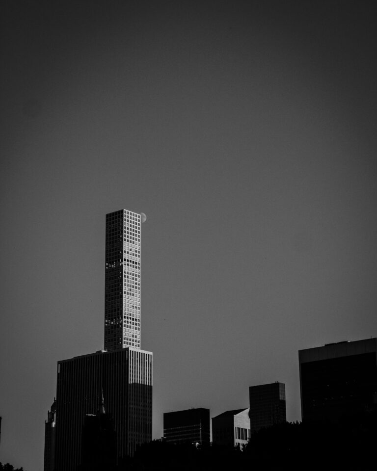 Black-and-white cityscape featuring a tall, grid-pattern skyscraper with a faint moon nearby in the sky and other dark buildings at street level.