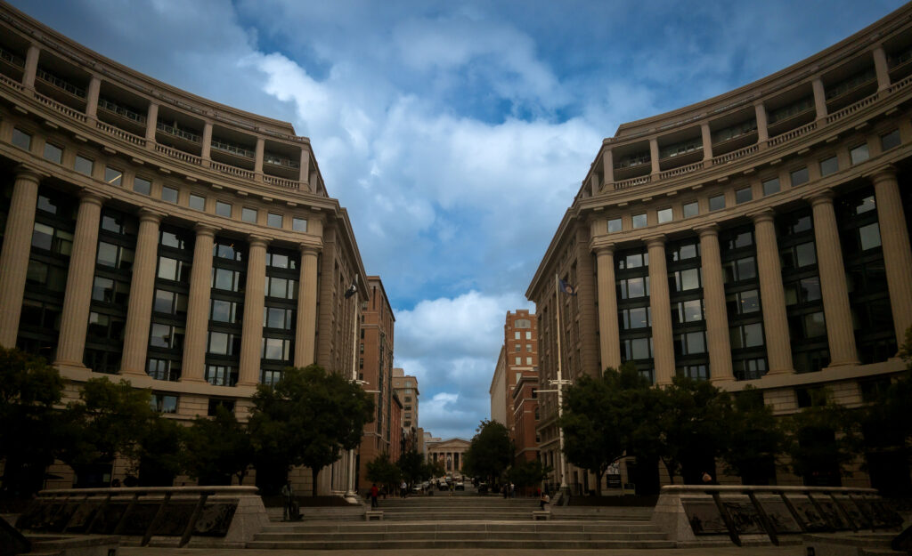 City street framed by curved neoclassical buildings with tall columns and large windows, converging toward a blue, cloud-filled sky.