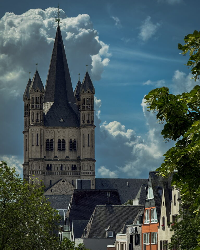 Tall Gothic church tower with spires rising above a row of townhouses against a blue sky with clouds and green trees nearby.