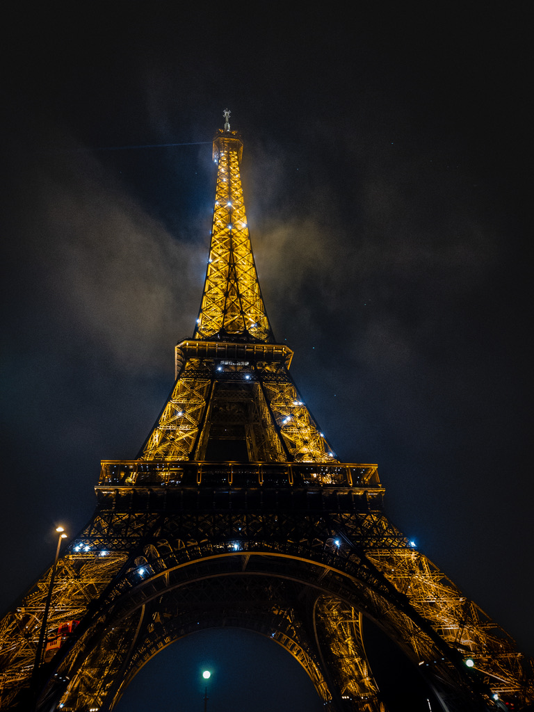 Eiffel Tower at night, illuminated in warm gold lights, seen from the base with a dark cloudy sky behind it.