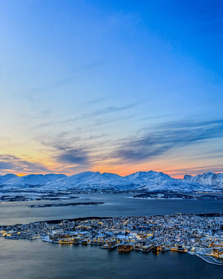 Aerial view of a snow-covered coastal town at sunset, with mountains in the distance and a calm, icy harbor.