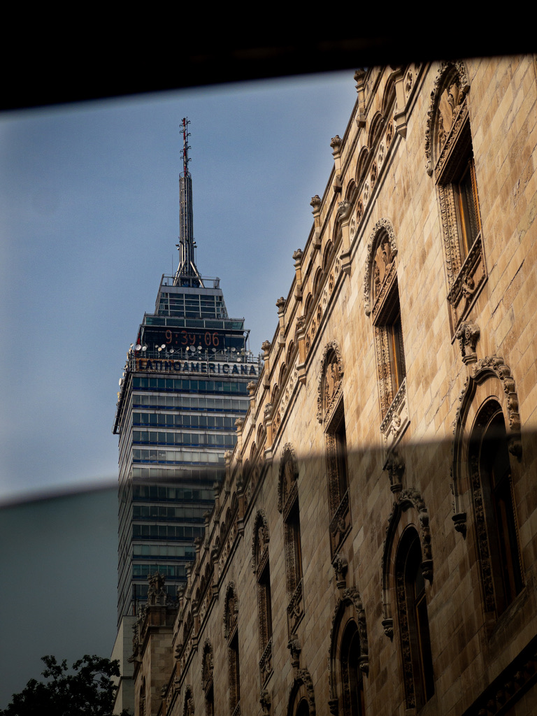 View of a historic stone building with arched windows on the right, beside a tall modern skyscraper with an antenna, seen through a dark window frame.
