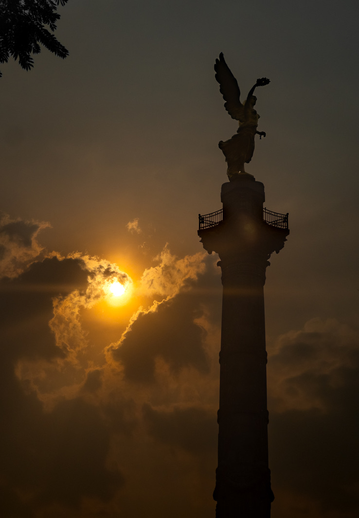 Silhouette of a winged statue atop a tall column against a sunset sky.